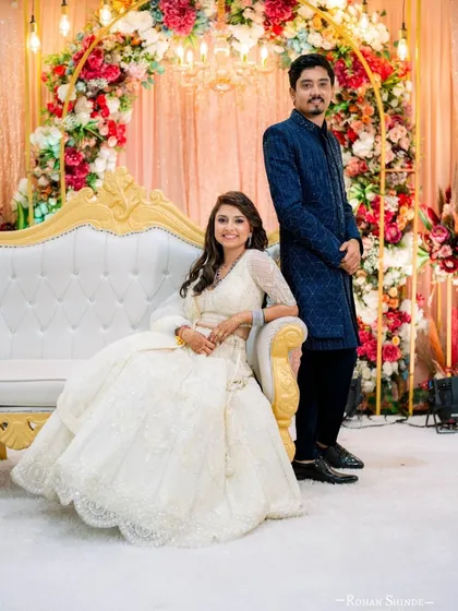 The same beautiful couple posing on their reception stage. The full view showcases the elegant flare of her white lehenga and the sharp tailoring of his blue bandhgala, a perfectly styled pair for their special day.