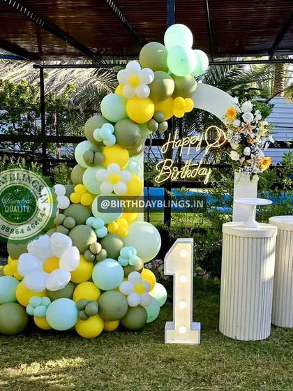An outdoor daisy-themed first birthday party. The balloon arch in shades of green and yellow, combined with white plinths and a light-up '1', creates a beautiful garden party feel.