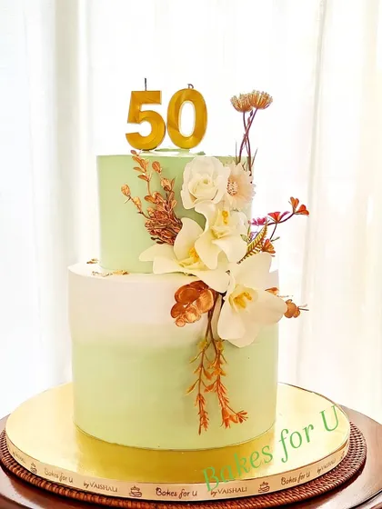 A sophisticated two-tier 50th birthday cake with a soft green ombre effect. The cake is decorated with an arrangement of beautiful white and gold dried flowers.