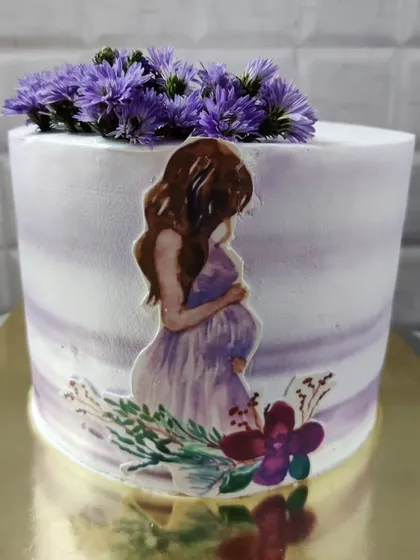 A beautiful baby shower cake for a mom-to-be. The design features a watercolor effect and is topped with fresh purple flowers.