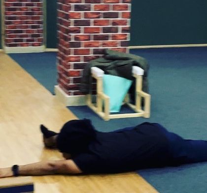 A student practices a preparatory pose for Hanumanasana (the splits), lying flat on the floor to stretch the hip flexors.