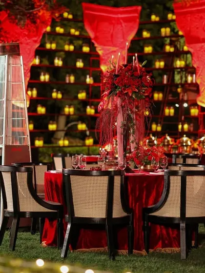 An outdoor dining table set for a sangeet, featuring a lush red floral centerpiece and surrounded by the warm glow of a candle wall in the background.