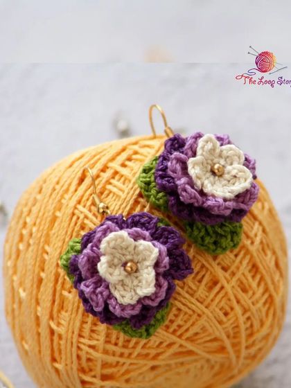 A pair of purple pansy-style crochet earrings resting on a ball of yarn, showing the vibrant colors I love to work with.