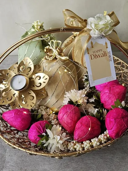 A golden wire basket filled with bright pink decorative lotuses, potlis, and a floral T-light holder. The fairy lights add a magical touch, making this a truly festive and joyful Diwali hamper.