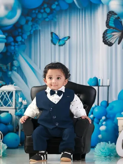 A regal shot of the birthday boy sitting on a mini armchair, looking dapper against the elaborate blue balloon arch.