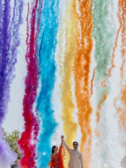 A spectacular moment of celebration at a rust-orange themed Haldi, with rainbow-colored smoke bombs creating a stunning visual against the sky.