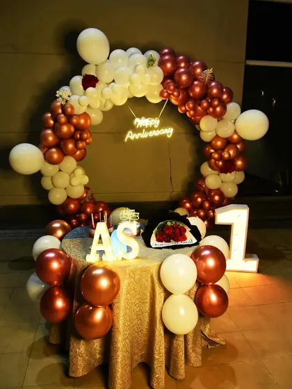 A romantic first-anniversary setup. A circular balloon arch in chrome rose gold and white frames a table set for two, with initial and number props.