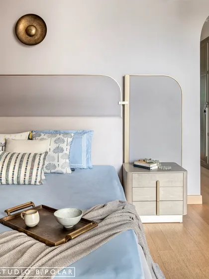The bed is styled with soft blue linens and a variety of cushions, adding layers of comfort and subtle color to the neutral room. A brass wall accent adds a touch of metallic warmth.