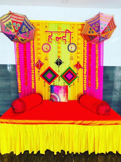 A colourful Haldi seating area with a bright red divan, set against a backdrop of pink and yellow drapes and decorative umbrellas.