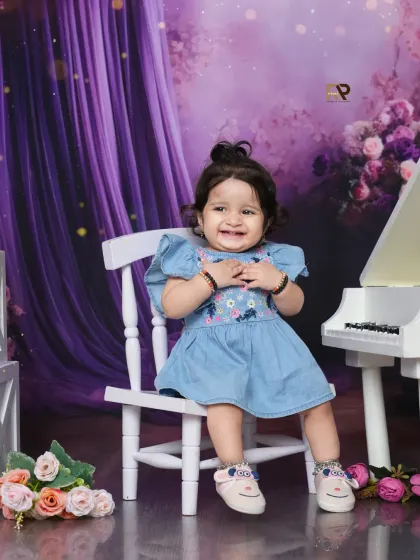 A baby girl in a denim dress sits on a white chair next to a piano in a purple floral wonderland. Her happy smile lights up this magical and harmonious scene.