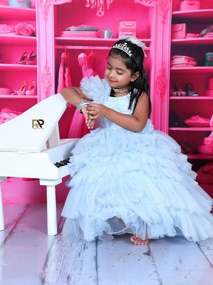 A young girl in a blue princess gown plays with her bracelet next to a miniature piano. The all-pink closet backdrop creates a stylish and glamorous setting fit for royalty.