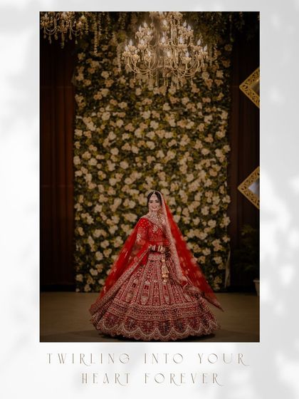 The bride twirls in her red lehenga against a wall of white flowers and under a grand chandelier, a perfect bridal portrait.