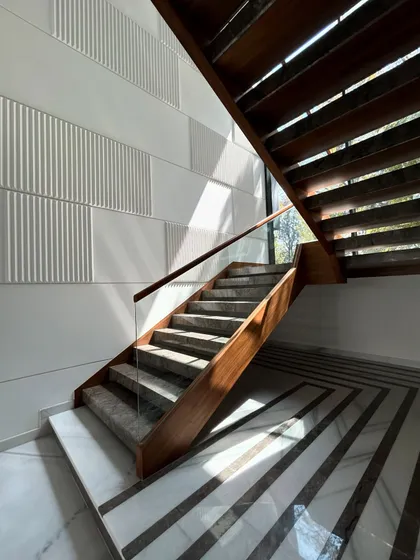 The modern staircase at Gulmohar Farmhouse features stone steps, a glass railing, and a warm wood structure. The striped marble floor at the base adds another layer of geometric detail.