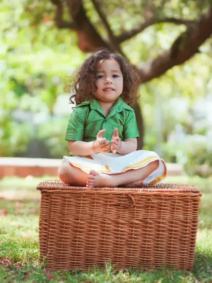 A full-body shot of a toddler in a traditional South Indian outfit, sitting on a wicker basket during an outdoor shoot. The natural setting is perfect for cultural portraits.