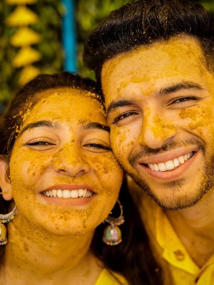 A happy, close-up portrait of a couple during their haldi ceremony, their faces covered in turmeric paste, smiling brightly for the camera.