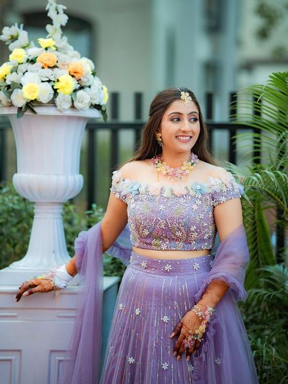 Dreamy floral and purple hues for a Haldi ceremony. This look is soft, romantic, and perfectly coordinated with her beautiful off-shoulder lehenga.