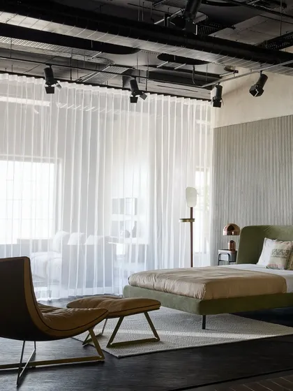 A closer look at the bedroom setup, focusing on the olive green bed and the minimalist lounge chair and ottoman. The track lighting provides focused illumination, typical of a gallery setting.