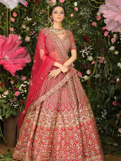 A full-length bridal fashion shot of a model in an elaborate red lehenga, set against a beautiful wall of flowers. This image is ideal for bridal couture lookbooks and campaign photography.