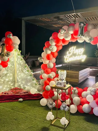 This cozy and stylish terrace setup is perfect for a first anniversary. It features a beautiful red, white, and silver balloon arch, a lit-up teepee for a romantic seating area, and a custom neon sign to celebrate your special day.
