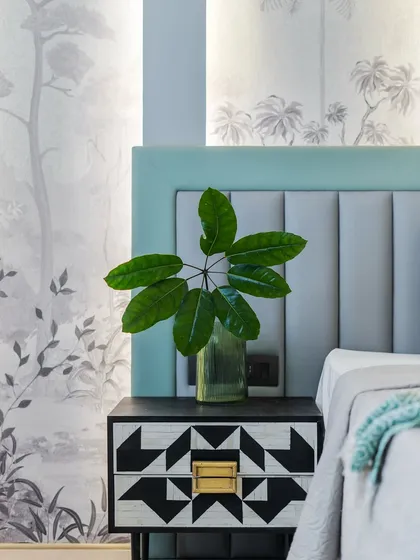 A detail shot of a bedside table with a striking black and white geometric pattern. The soft green headboard and delicate grey wallpaper create a calm and soothing backdrop, while a single green leaf adds a touch of nature.