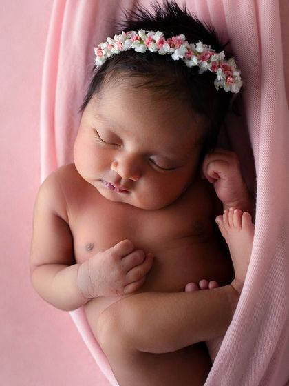 A three-week-old baby girl suspended in a soft pink sling. The gentle color and womb-like pose create a feeling of warmth, safety, and innocence.