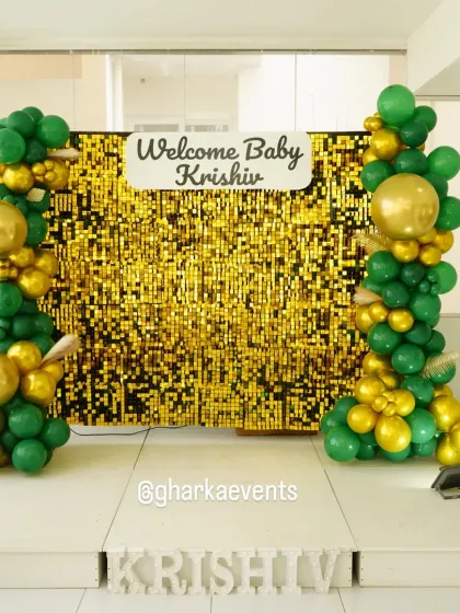A glamorous "Welcome Baby" setup with a gold sequin wall. The shimmering backdrop is flanked by balloon columns in dark green and gold, creating a rich and celebratory look.