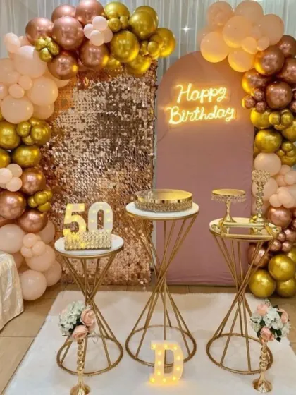 A glamorous 50th birthday celebration with a rose gold sequin wall. The setup includes metallic balloon arrangements, marquee numbers, and elegant gold plinths.