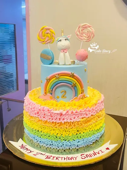 A vibrant two-tier unicorn cake with rainbow-colored ruffles. The top tier features a cute unicorn, lollipops, and a rainbow, making it a sweet fantasy for a 2nd birthday.