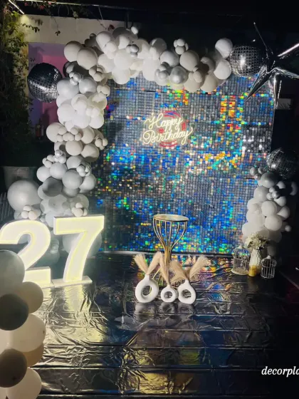 A full view of a glamorous silver-themed birthday party. The combination of a reflective floor, a shimmering sequin wall, and an arch of silver and white balloons creates an immersive and luxurious experience for a special 27th birthday.