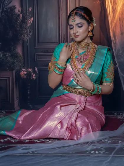 A full-length seated portrait in a stunning teal and pink silk saree. The makeup is soft and romantic, matching the mood of the shot.