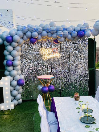 A first anniversary party on a terrace, highlighted by a silver sequin backdrop, a purple and silver balloon arch, and a large light-up number '1'.
