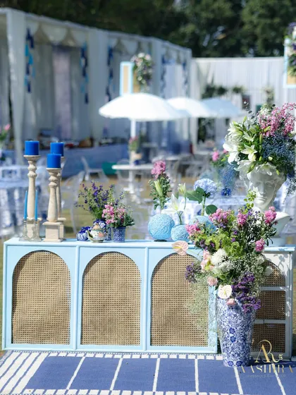 A charming console table setup with a rattan front, adorned with blue candles, and surrounded by beautiful arrangements of blue and purple florals in patterned vases.