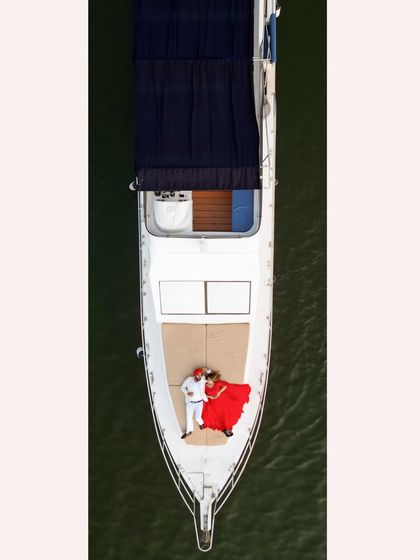 A breathtaking aerial drone shot of the couple lying on the bow of a yacht. The contrast of her red dress against the green water creates a stunning, artistic image.