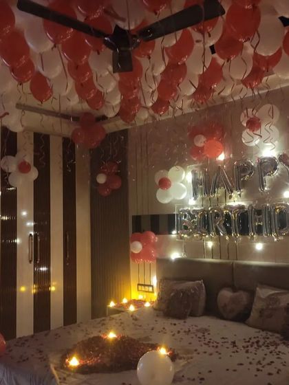 The final look of a romantic birthday room decoration, with hundreds of balloons on the ceiling, "Happy Birthday" foil letters, and the bed decorated with candles and rose petals.