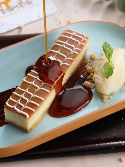 An action shot that adds a touch of luxury. The caramel being poured over the cheesecake creates a mouth-watering moment, highlighting the dessert's rich texture and flavor profile for a restaurant's dessert menu.