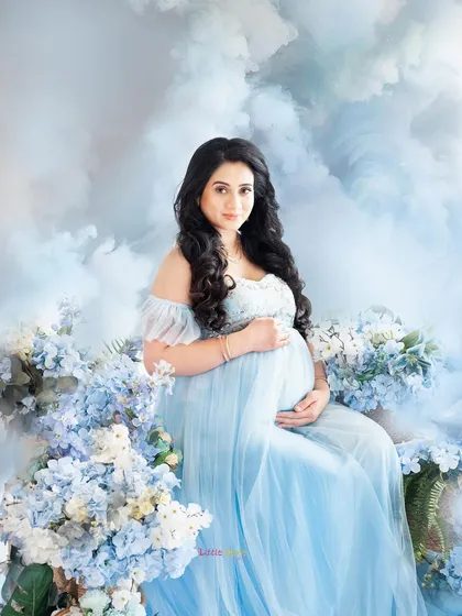 A beautiful, fantasy-themed portrait of a mother-to-be. She is seated in a light blue gown amidst a bed of blue flowers and soft, cloud-like textures, creating a serene and magical atmosphere.