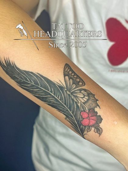 A composite tattoo of a large feather, a butterfly, and a small red flower, blending different elements of nature.