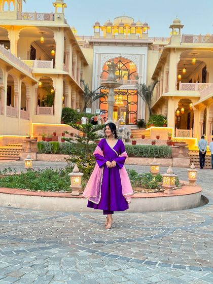 A full view of the purple anarkali suit in a stunning palace-like setting. I can help you choose outfits that not only suit you but also complement your event's location.