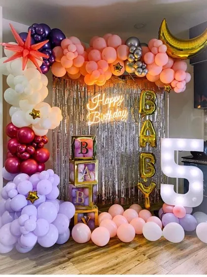 A colorful 5th birthday party setup with a silver shimmer backdrop, 'BABY' balloon boxes, and a large '5' marquee light.
