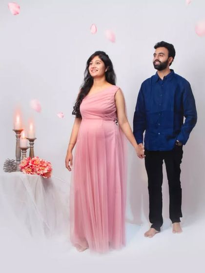 A romantic studio setup with a couple holding hands. The mom-to-be wears a beautiful one-shoulder pink gown, and the scene is accented with candles and falling petals for a dreamy effect.
