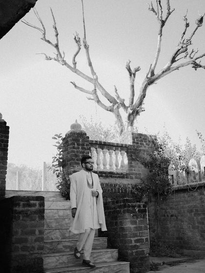 A striking black and white solo portrait of the groom on a stone staircase. The dramatic silhouette of the bare tree behind him adds an artistic, high-contrast element.