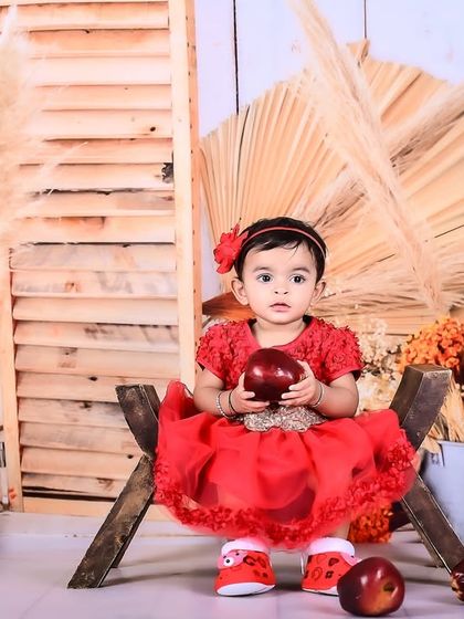 A baby girl in a red dress sitting in a rustic setup with apples and pampas grass.