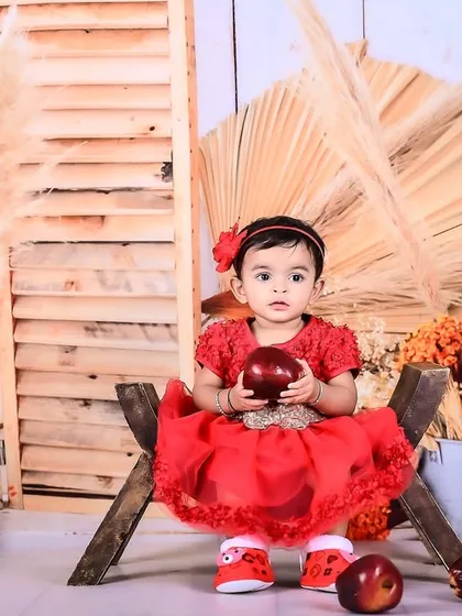 A baby girl in a red dress sitting in a rustic setup with apples and pampas grass.