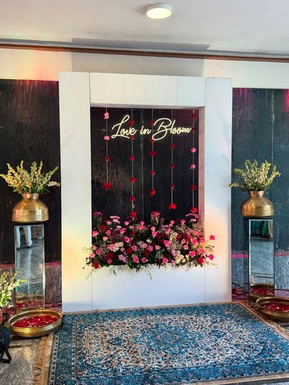 A "Love in Bloom" neon sign set within a white frame and surrounded by flowers, creating a modern and romantic photo booth for the couple and guests.
