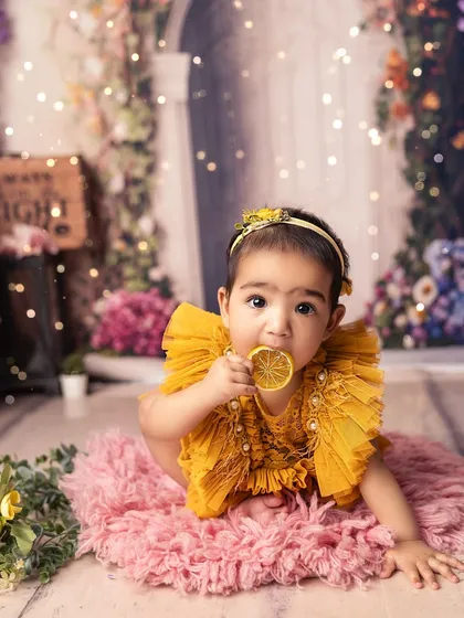 A little sunshine tasting a slice of sweetness. The combination of the floral background, her bright yellow dress, and the lemon prop creates a vibrant and joyful image.