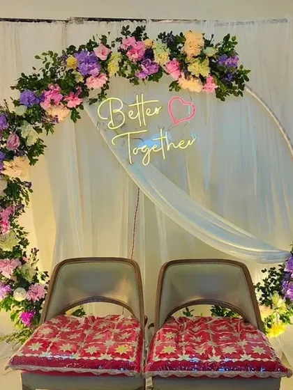 A beautiful floral ring backdrop for an intimate engagement or sangeet. The arch is densely decorated with a mix of colorful artificial flowers and features a "Better Together" neon sign, adding a romantic glow to the couple's seating area.