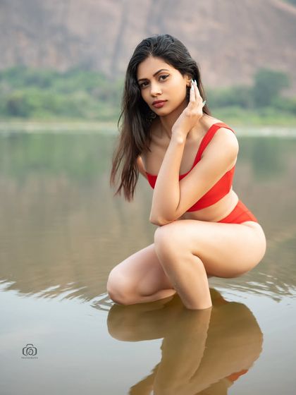 A close-up portrait of a model in a red swimsuit, kneeling in the water. The vibrant red color stands out beautifully against the natural tones of the lake and the surrounding landscape.