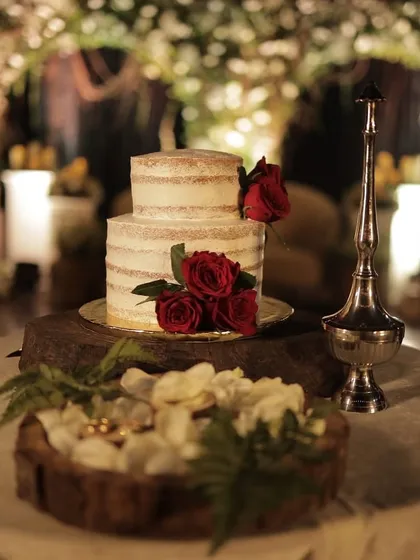 A sweet ending to a new beginning. This elegant two-tiered naked wedding cake, adorned with fresh red roses, sits on a rustic wooden stand, perfectly capturing a blend of classic romance and modern simplicity.