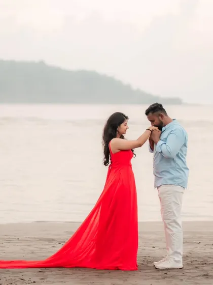 A tender moment on the beach. The groom-to-be kisses his partner's hand, her red trail gown pooling elegantly on the sand.