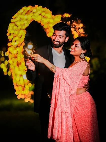 A magical night pre-wedding photo where the couple holds sparklers, standing before a heart-shaped arch of golden lights that creates a beautiful bokeh effect.